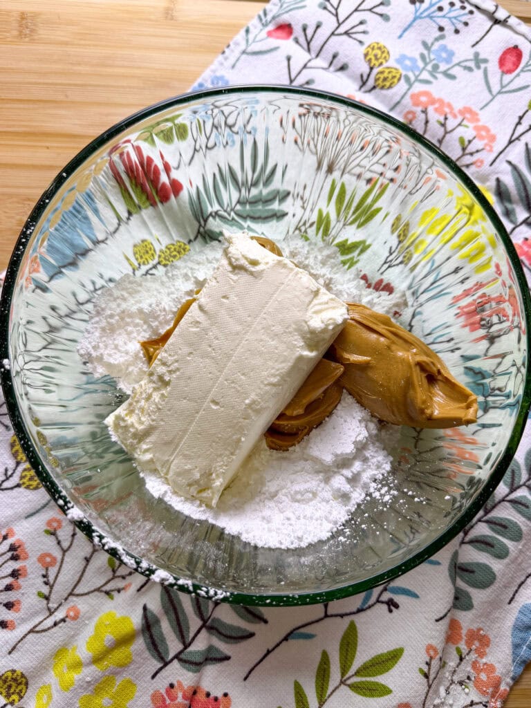 A bowl containing cream cheese, peanut butter, and powdered sugar sits on a floral-patterned cloth—perfect for creating a delicious Chocolate Peanut Butter Tiramisu—on a wooden surface.