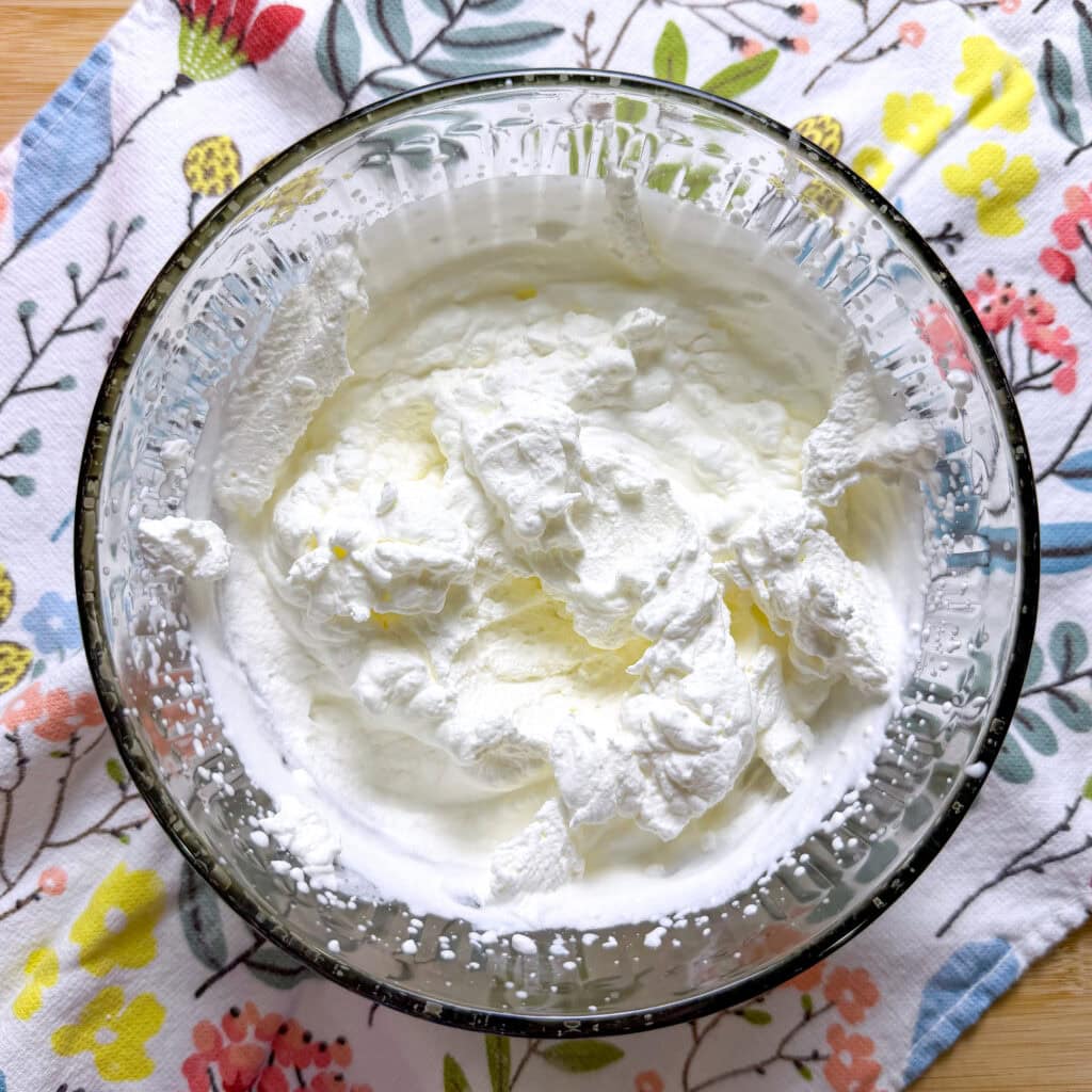 A glass bowl filled with fluffy whipped cream sits on a colorful, floral-patterned cloth atop a light wooden surface.