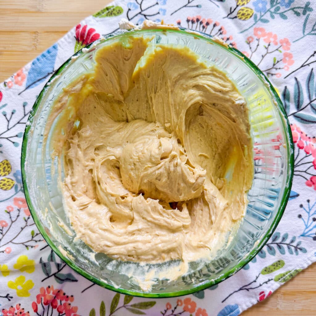 A glass bowl filled with creamy, light brown cake batter sits on a colorful, floral-patterned cloth atop a wooden surface.