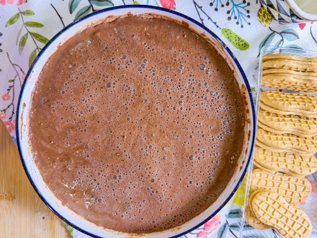 A bowl filled with frothy chocolate mixture sits on a floral tablecloth, accompanied by a row of peanut-shaped cookies—perfect for assembling a delightful Chocolate Peanut Butter Tiramisu.