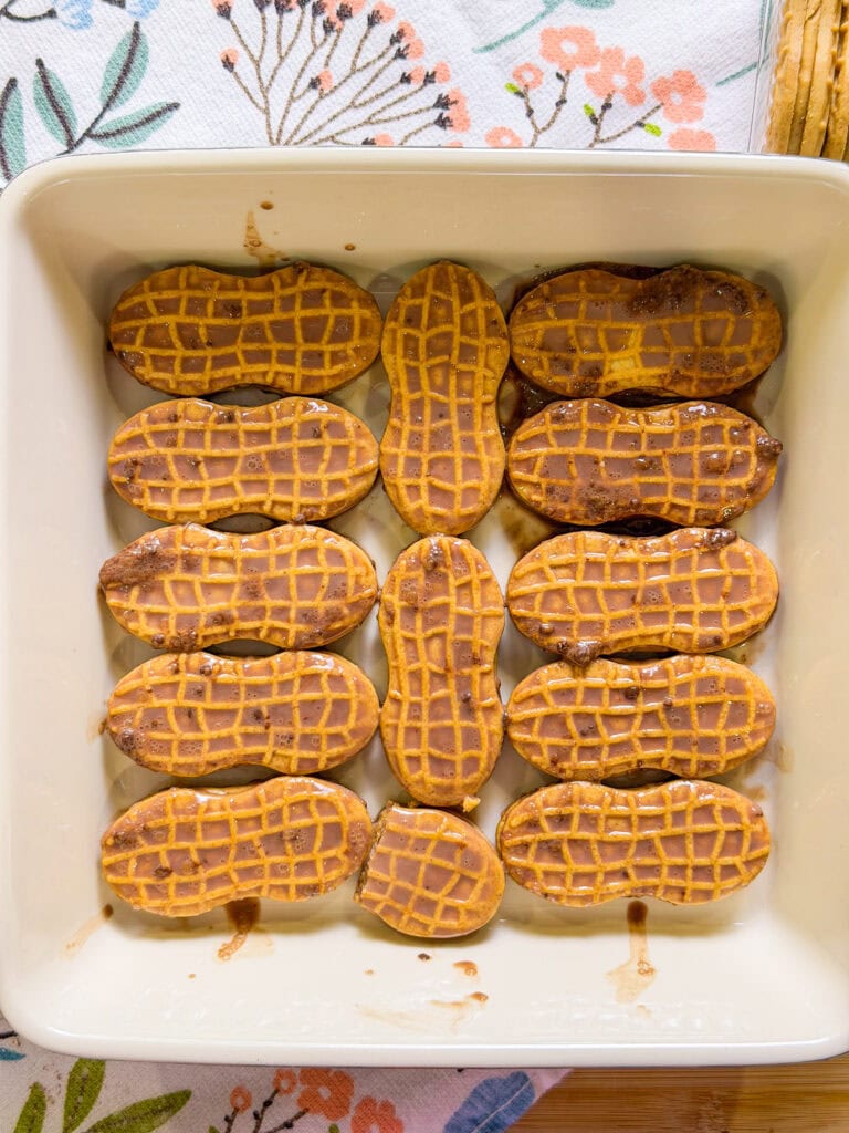 A square baking dish filled with a single layer of Nutter Butter cookies, some with chocolate and cream residue, on a floral-patterned cloth background.
