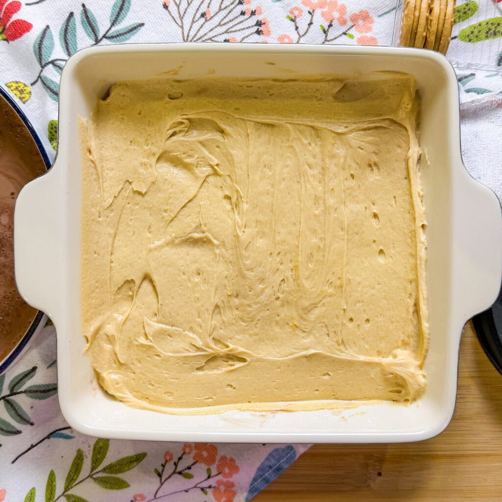 A square white baking dish filled with smooth, light brown batter sits on a floral-patterned cloth, ready to be baked. A wooden spoon and partial bowls are visible nearby on the table.