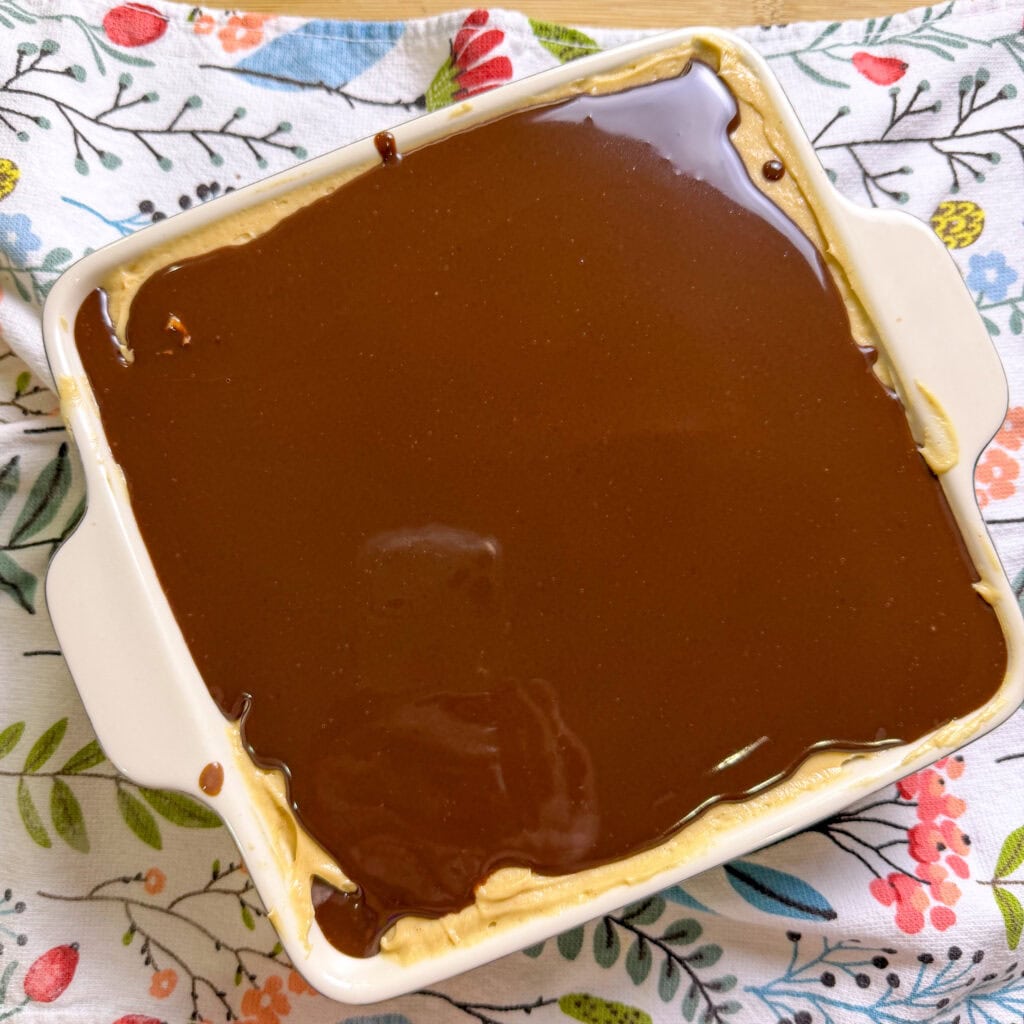 A square white dish filled with a dessert topped with a smooth layer of chocolate, sitting on a colorful floral-patterned cloth.