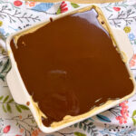 A square white dish filled with a dessert topped with a smooth layer of chocolate, sitting on a colorful floral-patterned cloth.