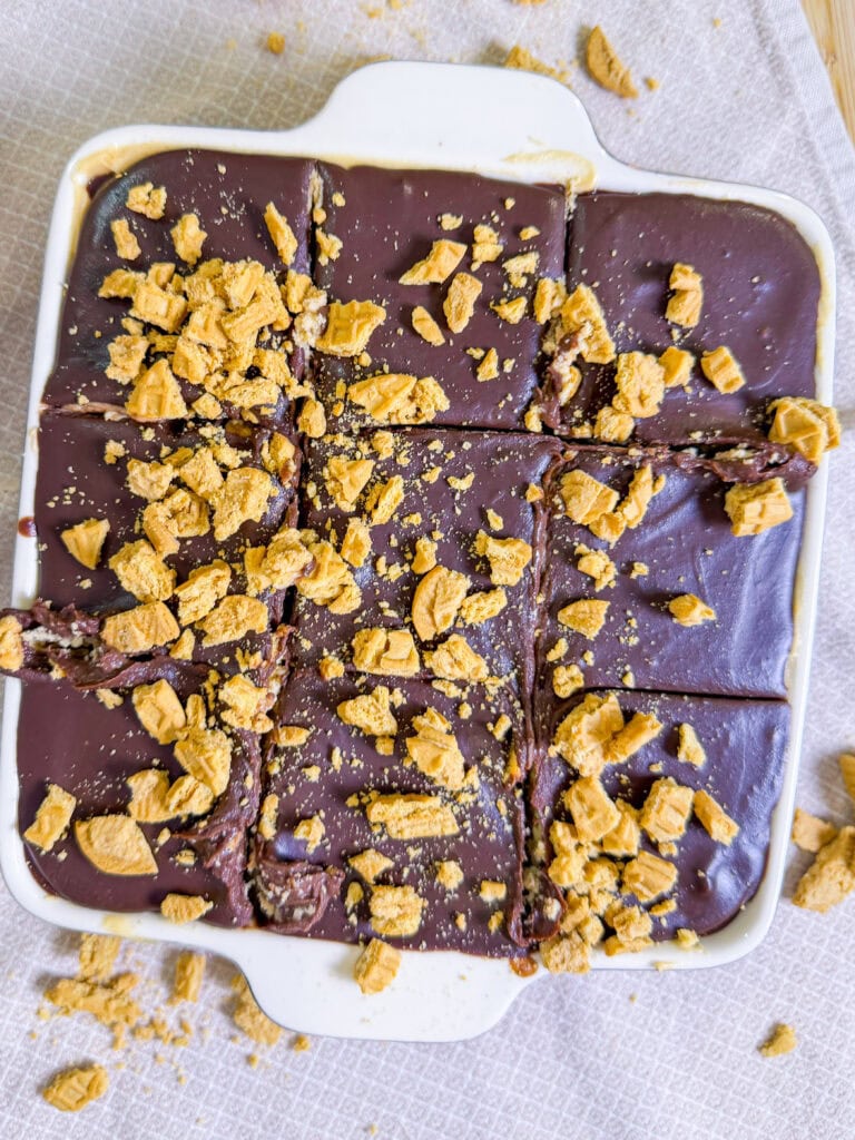A square white dish filled with Chocolate Peanut Butter Tiramisu bars, sliced into nine pieces and topped with crumbled golden cookies. The dish sits on a light textured cloth, with some crumbs scattered around.