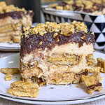 A close-up of a layered dessert with cream, cookies, and a chocolate topping, garnished with crumbled cookies on a white plate. More dessert and a patterned bowl are blurred in the background.