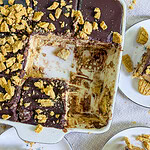 A partially eaten chocolate and graham cracker dessert in a rectangular baking dish, with three plates nearby holding individual servings topped with crumbled graham crackers. Crumbs are scattered on the table.