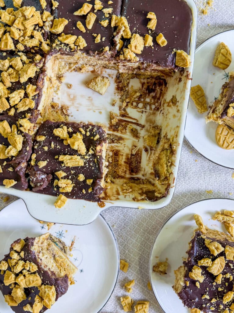 A partially eaten chocolate and graham cracker dessert in a rectangular baking dish, with three plates nearby holding individual servings topped with crumbled graham crackers. Crumbs are scattered on the table.