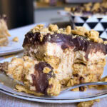 A close-up of a dessert made with layers of chocolate, creamy filling, and crushed cookies on a plate. A spoon with a piece of the dessert rests in the foreground. Another serving is visible in the background.