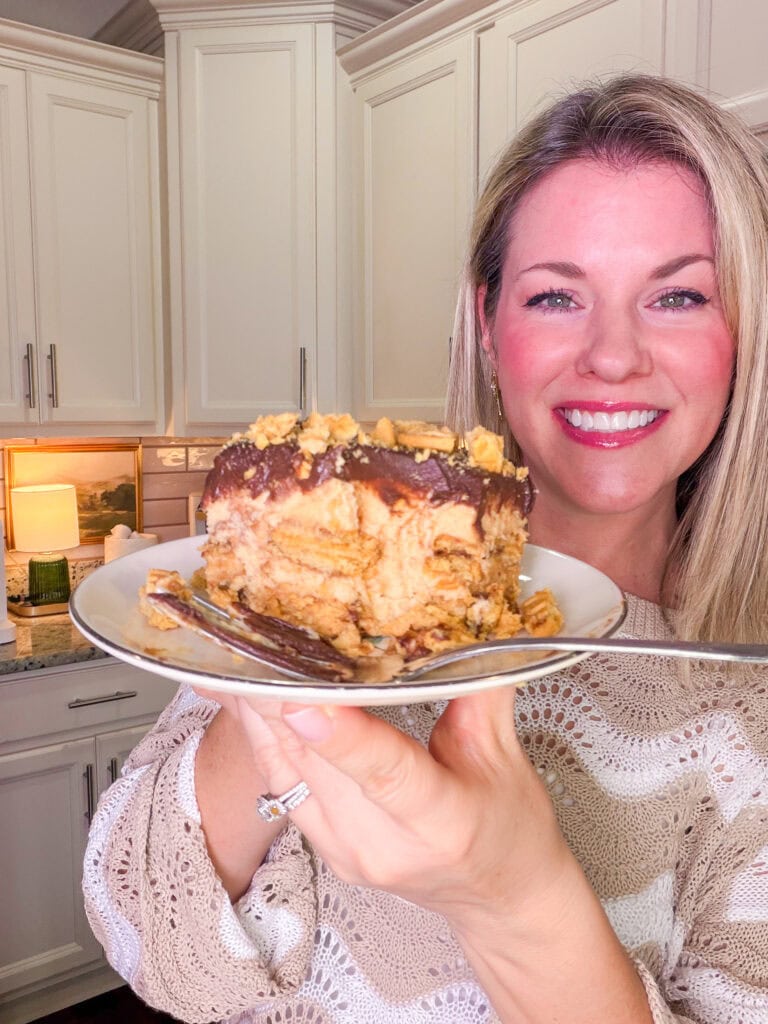 A smiling woman in a cream sweater holds up a plate with a large slice of layered chocolate and peanut butter dessert in a kitchen.