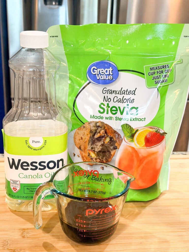 A bottle of Wesson canola oil, a bag of Great Value granulated stevia, and a glass measuring cup with a dark liquid sit on a wooden surface in a kitchen.