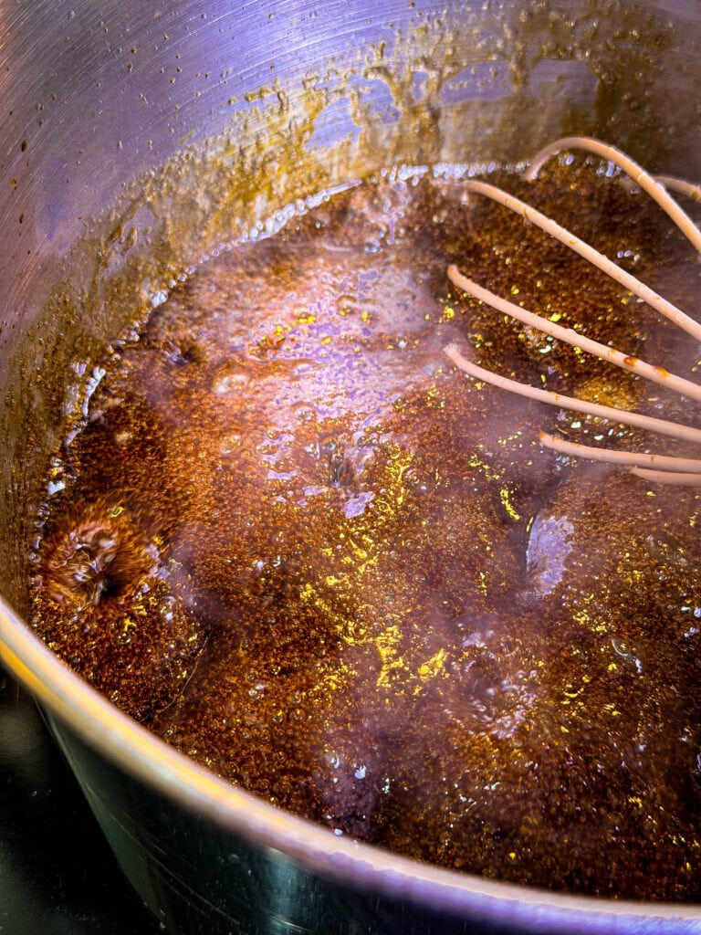 A pot of dark brown liquid is boiling with bubbles on the surface. A metal whisk is partially submerged in the mixture, and light reflects off the liquid inside the pot.