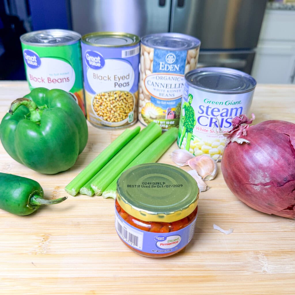 A wooden cutting board holds a green bell pepper, two celery stalks, a red onion, garlic, a jar of pimientos, and cans of black beans, black eyed peas, cannellini beans, and white shoepeg corn. A kitchen is in the background.