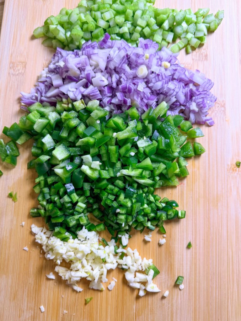 Chopped green bell peppers, red onions, celery, and garlic are arranged in rows on a wooden cutting board.