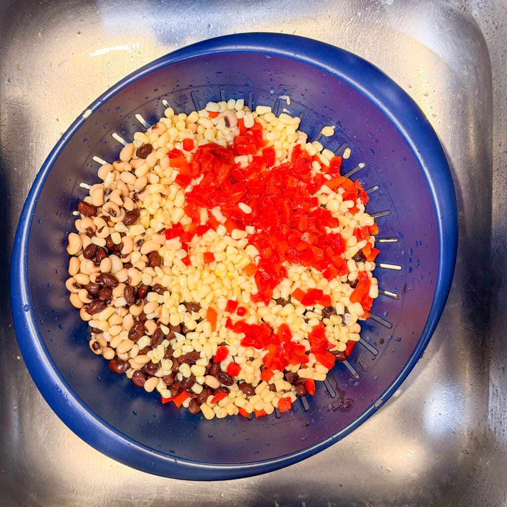 A blue colander in a sink contains a colorful mix of black beans, black-eyed peas, corn, and diced red bell peppers being rinsed.