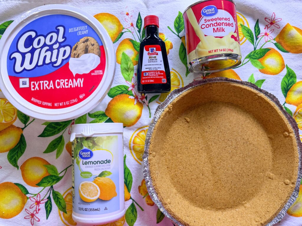Ingredients for a lemon pie arranged on a lemon-patterned tablecloth: Cool Whip, lemon concentrate, sweetened condensed milk, vanilla extract, and a graham cracker pie crust.