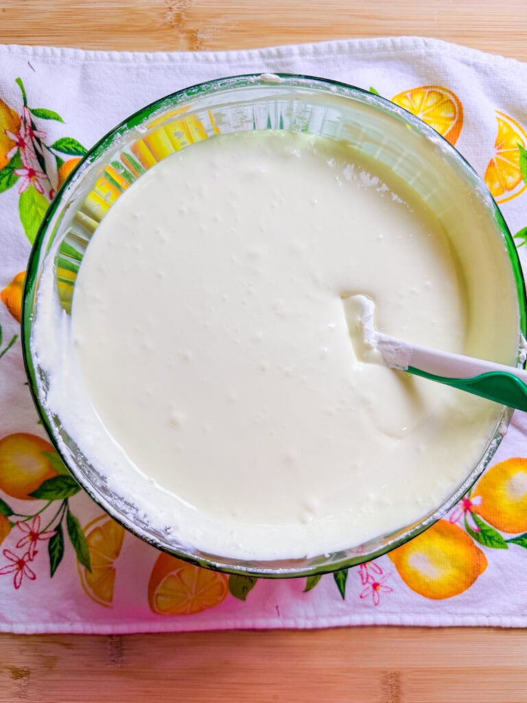 A glass bowl filled with smooth, creamy white batter and a green spatula sits on a decorative lemon-printed cloth on a wooden surface.