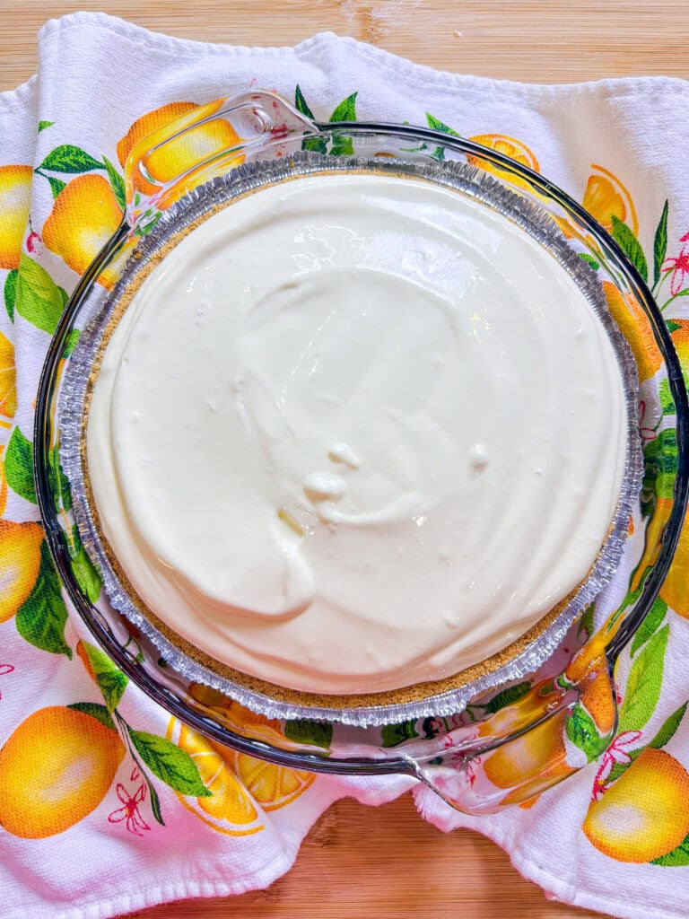 A glass pie dish filled with a creamy white pie, sitting on a colorful kitchen towel decorated with lemons and leaves, atop a light wooden surface.