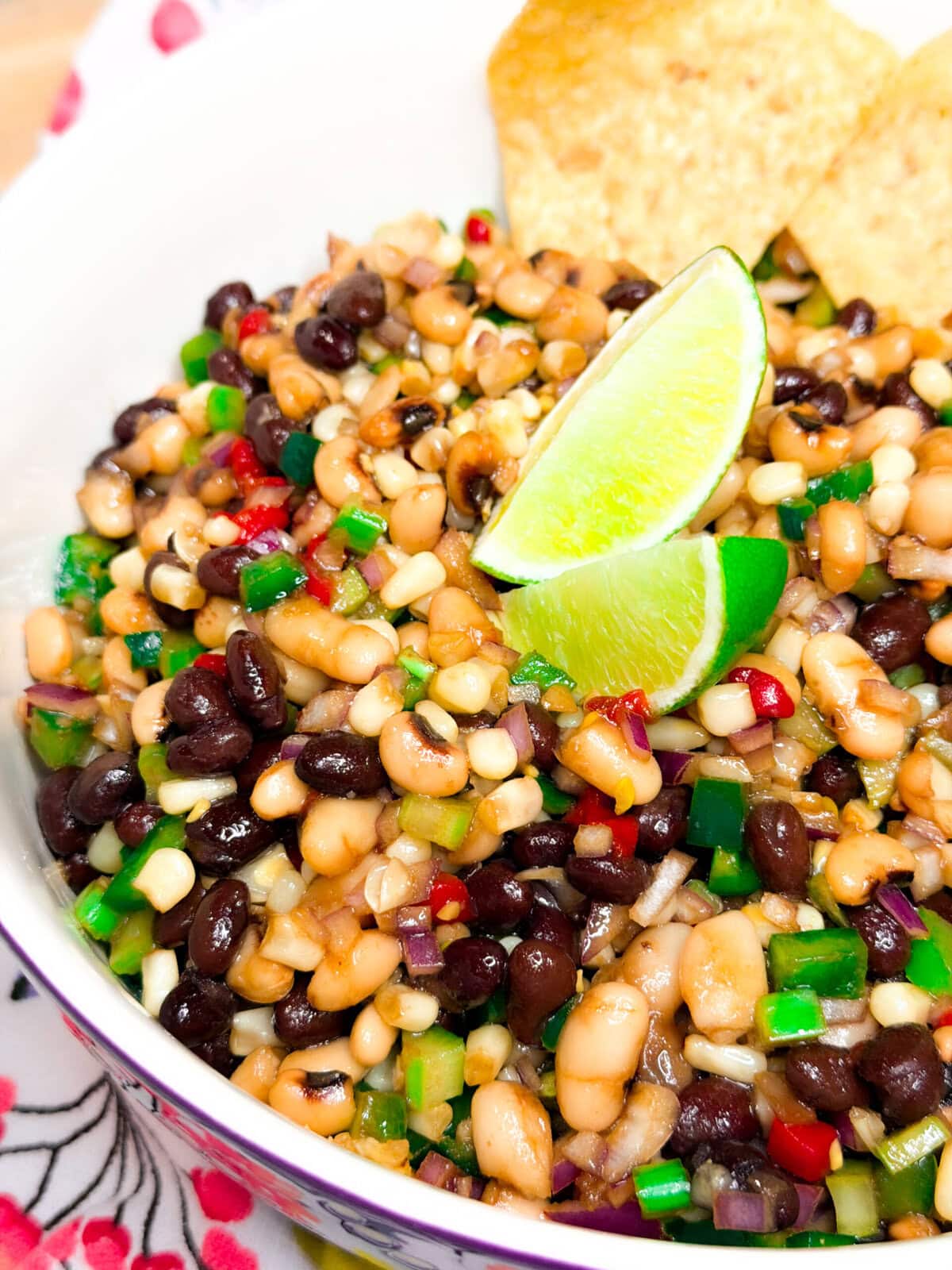A close-up of a colorful bean salad with black beans, white beans, chopped peppers, onions, and corn, garnished with two lime wedges and served with tortilla chips.