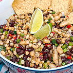 A bowl of bean salad with black beans, black-eyed peas, diced vegetables, and corn, garnished with lime wedges and three tortilla chips on the side.
