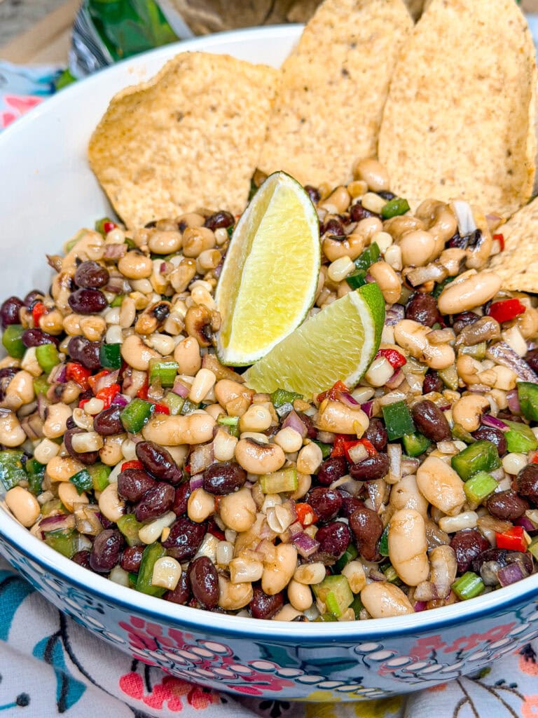 A bowl of bean salad with black beans, black-eyed peas, diced vegetables, and corn, garnished with lime wedges and three tortilla chips on the side.