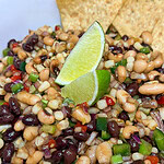 A close-up of a colorful bean salad with black beans, white beans, corn, chopped red onion, green onion, red peppers, and two lime wedges on top, served with tortilla chips in the background.