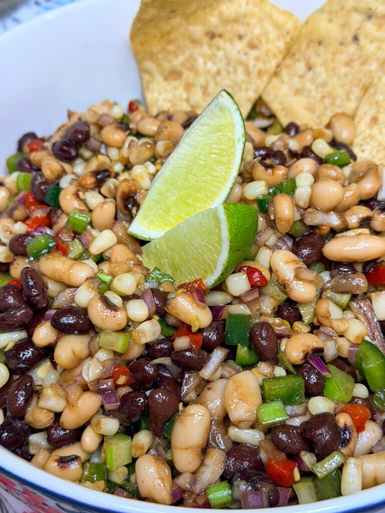 A close-up of a colorful bean salad with black beans, white beans, corn, chopped red onion, green onion, red peppers, and two lime wedges on top, served with tortilla chips in the background.