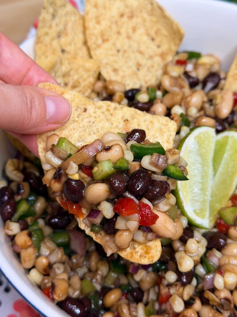 A hand holds a tortilla chip topped with a colorful bean salad made of black beans, black-eyed peas, corn, peppers, and onions. A lime wedge is visible in the bowl of salad in the background.