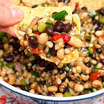 A hand holds a tortilla chip topped with a colorful bean salad containing black beans, black-eyed peas, peppers, corn, and red onion, with more salad and chips in a bowl below.