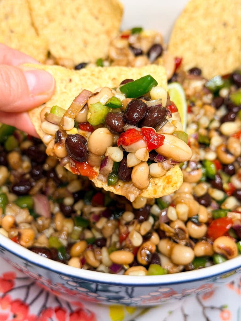 A hand holds a tortilla chip topped with a colorful bean salad containing black beans, black-eyed peas, peppers, corn, and red onion, with more salad and chips in a bowl below.