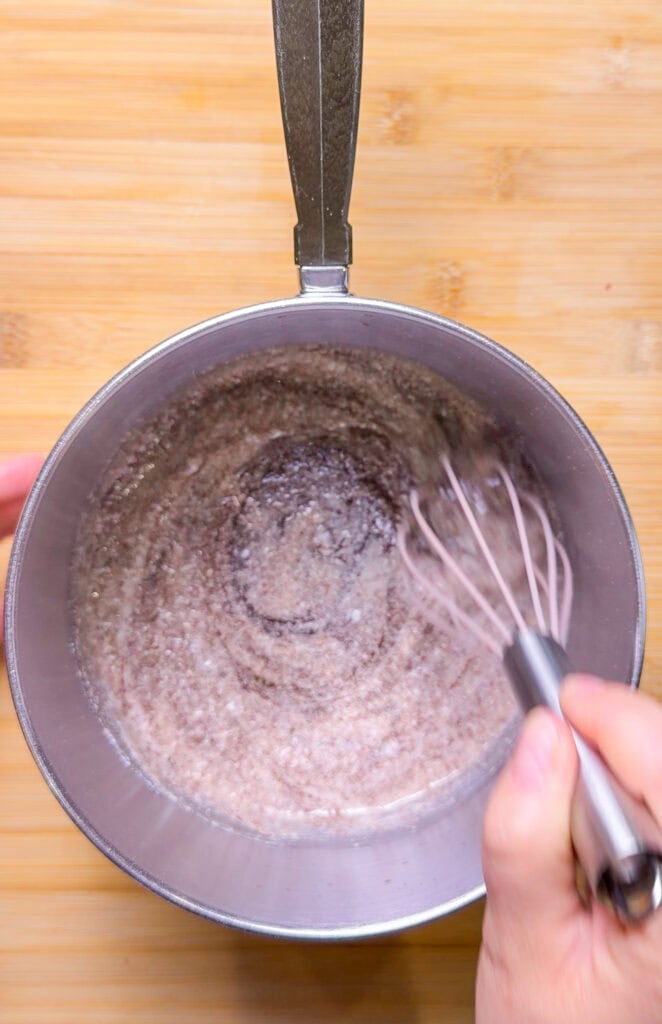 A person is whisking a mixture of dry ingredients in a metal bowl on a wooden surface. The photo is taken from above, showing the motion of the whisk stirring the contents.