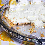 A creamy lemon pie with a graham cracker crust, topped with whipped cream and lemon slices, sits in a glass pie dish with one large slice missing. The background features a marble pattern and lemon-themed tablecloth.