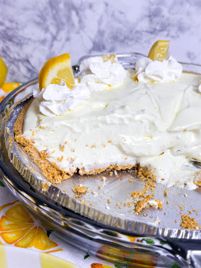 A creamy lemon pie with a graham cracker crust, topped with whipped cream and lemon slices, sits in a glass pie dish with one large slice missing. The background features a marble pattern and lemon-themed tablecloth.