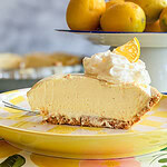 A slice of creamy lemon pie with whipped cream and a lemon wedge sits on a yellow plate. In the background, a bowl of fresh lemons is displayed on a white cake stand.