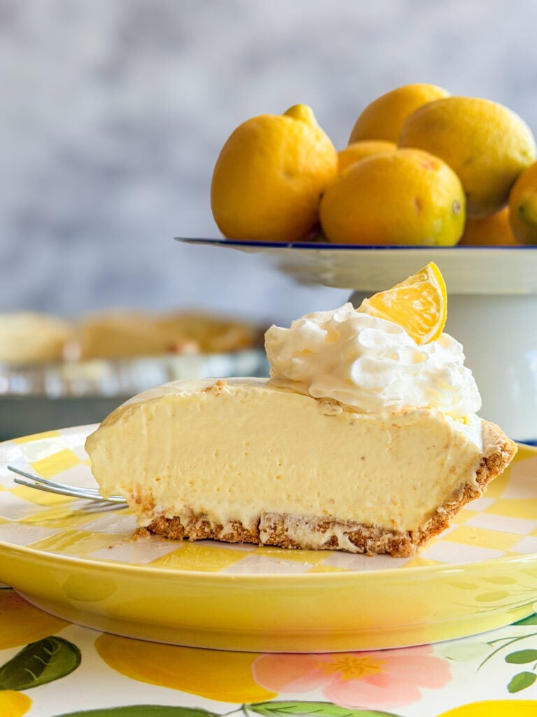 A slice of creamy lemon pie with whipped cream and a lemon wedge sits on a yellow plate. In the background, a bowl of fresh lemons is displayed on a white cake stand.