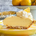 A slice of creamy lemon pie topped with whipped cream and a lemon wedge sits on a plate. In the background, theres a bowl filled with whole lemons. The scene is bright and colorful.