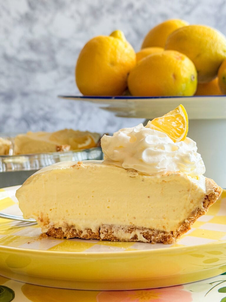 A slice of creamy lemon pie topped with whipped cream and a lemon wedge sits on a plate. In the background, theres a bowl filled with whole lemons. The scene is bright and colorful.