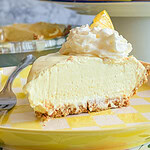 A slice of creamy lemon pie with whipped cream and a lemon wedge on top, served on a yellow checkered plate with a fork. In the background, there are whole lemons and a pie dish.