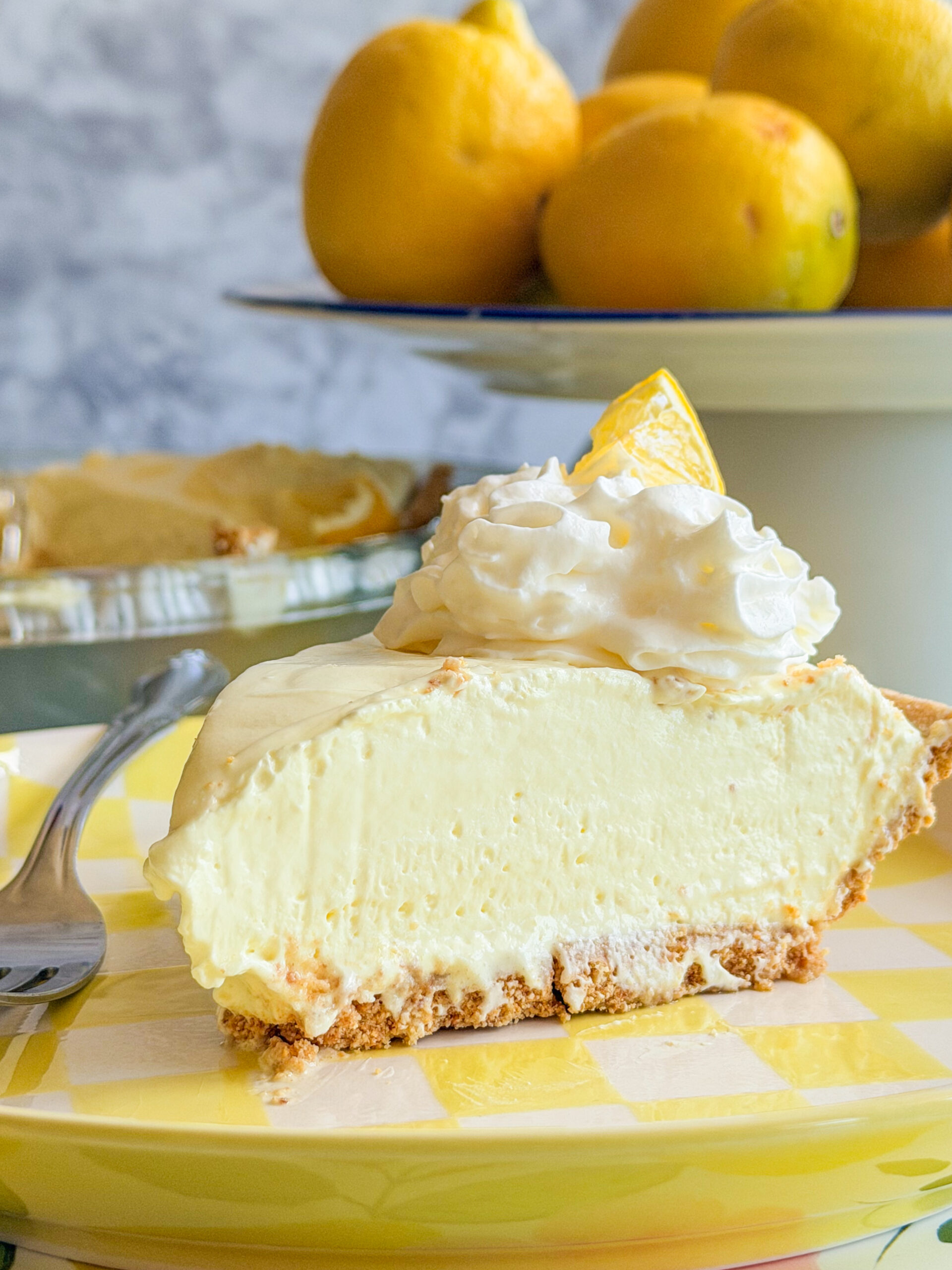 A slice of creamy lemon pie with whipped cream and a lemon wedge on top, served on a yellow checkered plate with a fork. In the background, there are whole lemons and a pie dish.