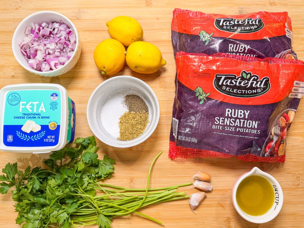 A wooden surface with bags of baby potatoes, a container of feta cheese, chopped red onion, lemons, garlic, fresh parsley, a bowl of spices, and a small cup of olive oil arranged neatly.