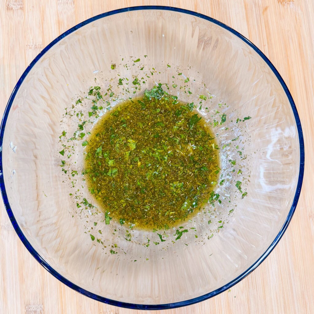 A clear glass bowl contains a small amount of green herb dressing or marinade, with finely chopped herbs and oil mixed together, placed on a light wooden surface.