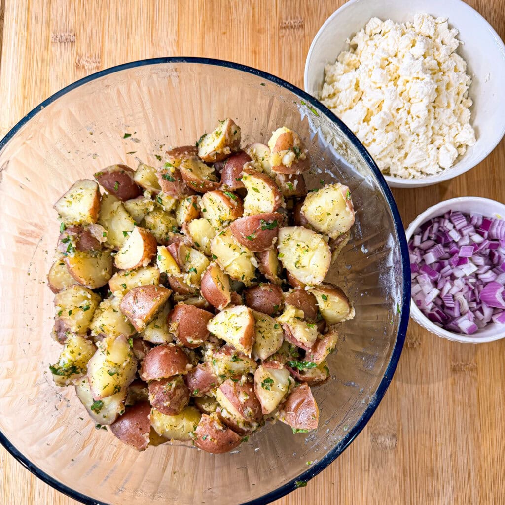 A glass bowl filled with herb-seasoned potato salad sits on a wooden surface, next to small bowls containing crumbled feta cheese and chopped red onions.