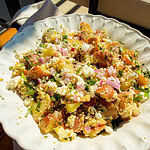 A white scalloped bowl filled with potato salad, featuring chunks of red potatoes, crumbled feta cheese, chopped red onions, and fresh herbs, set on a textured light-colored cloth in bright sunlight.