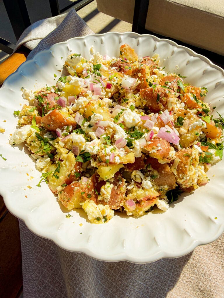A white scalloped bowl filled with potato salad, featuring chunks of red potatoes, crumbled feta cheese, chopped red onions, and fresh herbs, set on a textured light-colored cloth in bright sunlight.
