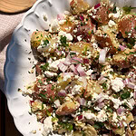 A white scalloped bowl filled with a potato salad made of red potatoes, crumbled feta cheese, chopped red onions, and fresh parsley. The dish is set on a wooden table with sunlight highlighting the ingredients.
