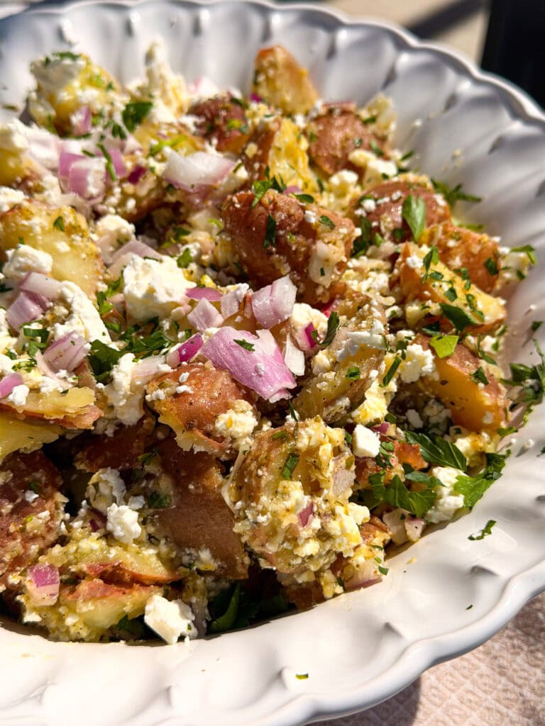 A close-up of a bowl filled with potato salad featuring red potatoes, chopped red onions, crumbled feta cheese, and fresh herbs, served in a white scalloped dish.
