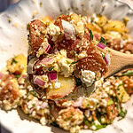 A wooden spoon holds a serving of potato salad with red potatoes, chopped red onion, crumbled feta cheese, and fresh herbs over a white bowl filled with more of the same salad.