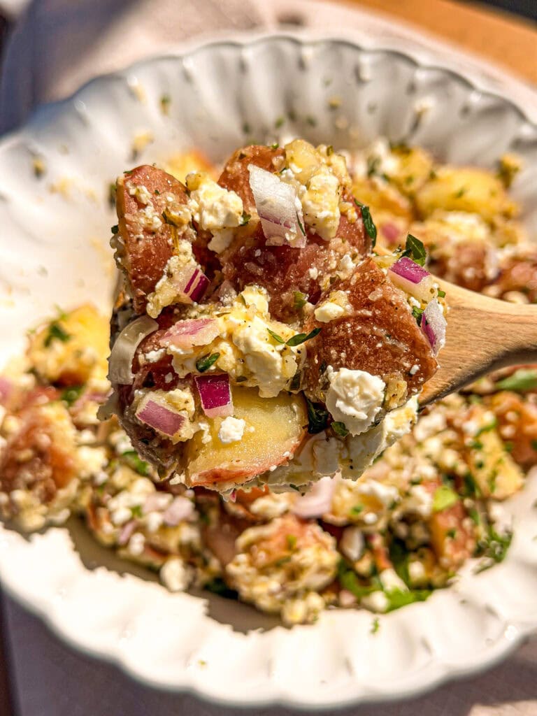 A wooden spoon holds a serving of potato salad with red potatoes, chopped red onion, crumbled feta cheese, and fresh herbs over a white bowl filled with more of the same salad.