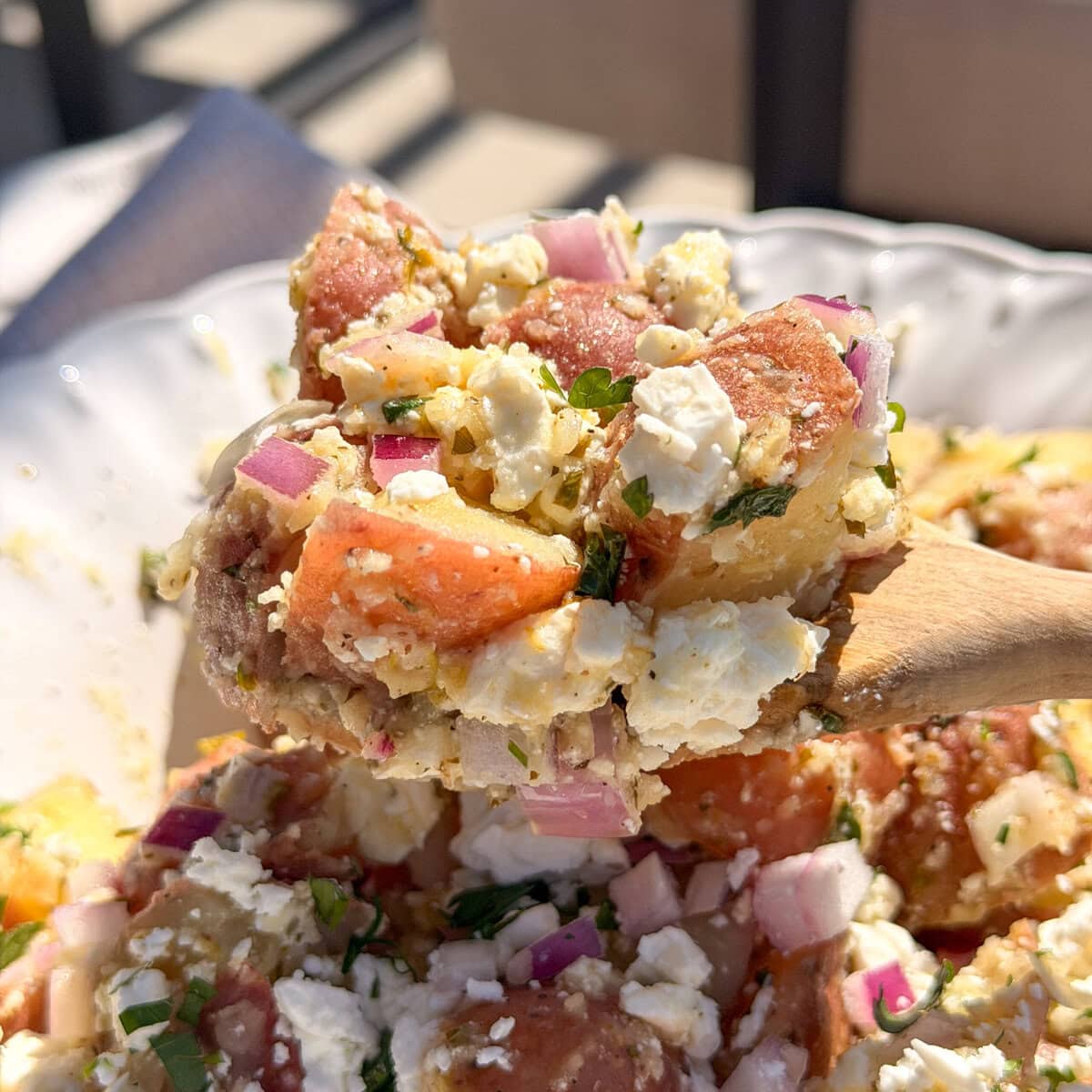A close-up of a wooden spoon holding a serving of potato salad with chopped red onions, feta cheese, herbs, and chunks of red potatoes in a white bowl. Sunlight shines on the dish.
