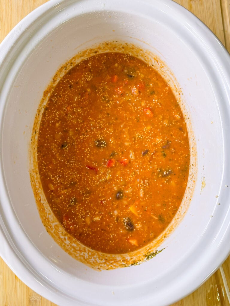 A white slow cooker filled with a thick, orange-red stew containing visible vegetables, beans, and grains, sitting on a wooden surface.